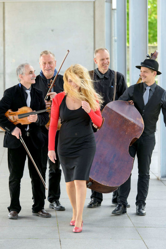 The String Company „Klezmer – Swing – Celtic“ Eine Frau, deren Haare im Wind wehen, schreitet voran. Im Hintergrund stehen vier Männer der Band mit Instrumenten in den Händen.