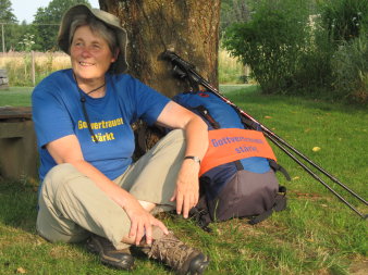 Foto: Brunhilde Schierl © Michaela Thoma Frau im Schneidersitz mit blauem T-Shirt mit der Aufschrift "Gottvertrauen stärkt" und Hut, rechts der Pilgerrucksack mit gleicher Aufschrift