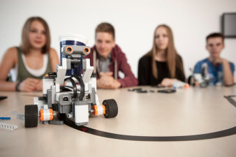 Foto: Angewandte Informatik für Kinder © Jens Hauspurg ein Robi im Vordergrund, im Hintergrund unscharf vier Jugendliche an einem Tisch sitzend