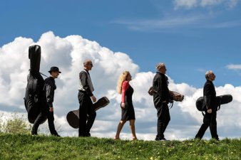 fünf Leute mit Instrumentenkoffern, in der Mitte eine Frau von links nach rechts auf Wiese vor Himmel laufend