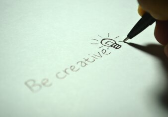 Ein Stift schreibt die englische Übersetzung von "Sei kreativ." auf ein Blatt Papier.