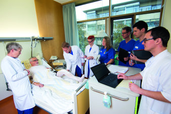 Foto: Unfallchirurgie, Orthophädie, Handchirurgie © Helios Kliniken GmbH einige weiß oder blau bekittelte Menschen am Patientenbett
