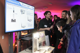 Foto: Kopfstand für 5G – Mobilfunk in Echtzeit © IMMS / Beate Hövelmans Ein Forscher des IMMS erklärt einer Gästegruppe den aus mehreren größeren Kompontenten bestehenden Aufbau zum inversen Pendel.
