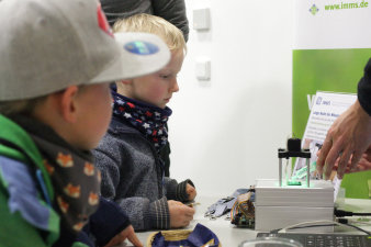 Foto: Krebs frühzeitig mit Mikroelektronik erkennen © IMMS / Beate Hövelmans Zwei Kinder schauen zu, wie Flüssigkeit aus einer Spritze direkt auf die Mikroelektronik eines brotdosengroßen Test