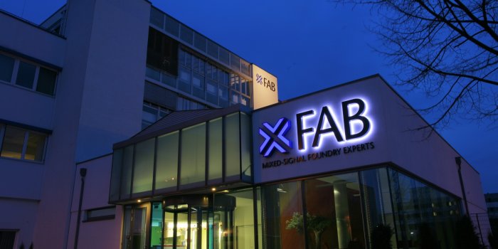 Foto: X-Fab Eingang bei Nacht © X-Fab / Beate Hövelmans Eingangsbereich zum Firmengebäude mit Glasfassade und leuchtendem Logo in der Dämmerung