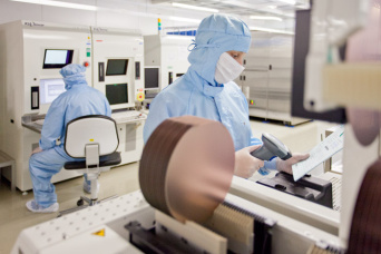 Foto: Mitarbeiter im MEMS-Center, einem speziellen Reinraum für MEMS-Technologien © Andreas Hultsch Mitarbeiter in blauen Anzügen mit Mundschutz im MEMS-Center, einem speziellen Reinraum für MEMS-Technologien. Im Vordergrund sind Siliziumscheiben auf einer Maschine zu sehen.