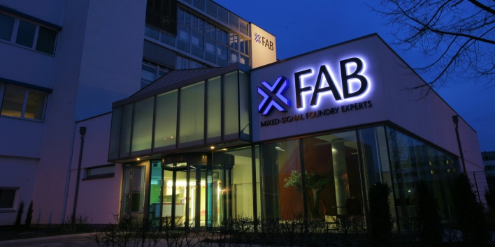 Foto: Eingang X-Fab © X-FAB / Beate Hövelmans Eingangsbereich zum Firmengebäude mit Glasfassade und leuchtendem Logo in der Dämmerung